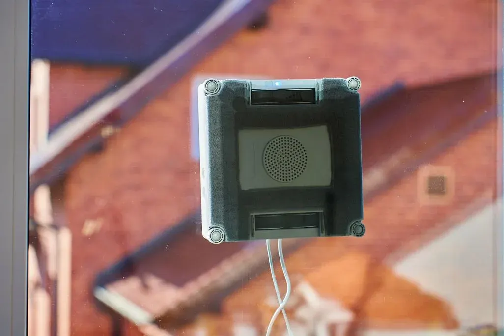 Die Unterseite eines Fensterputzoboters, der an der Scheibe haftet. Im Hintergrund sind Hausdächer zu sehen.