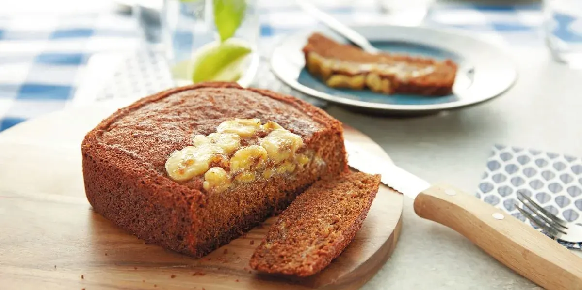 Ein angeschnittenes Bananenbrot liegt auf einem Brett, neben einer Scheibe, dahinter eine weitere Scheibe auf einem Teller.