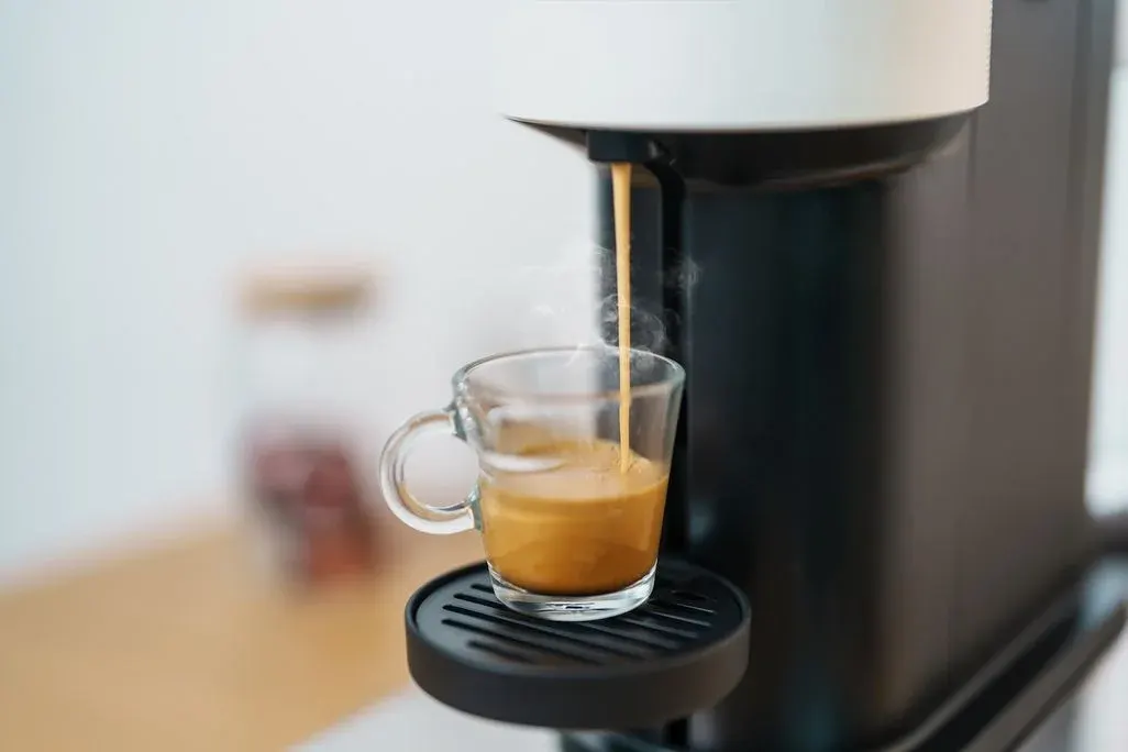 Nahaufnahme einer Kaffeemaschine, die frischen Espresso in eine gläserne Tasse laufen lässt; aufsteigender Dampf, unscharfer Küchenhintergrund.