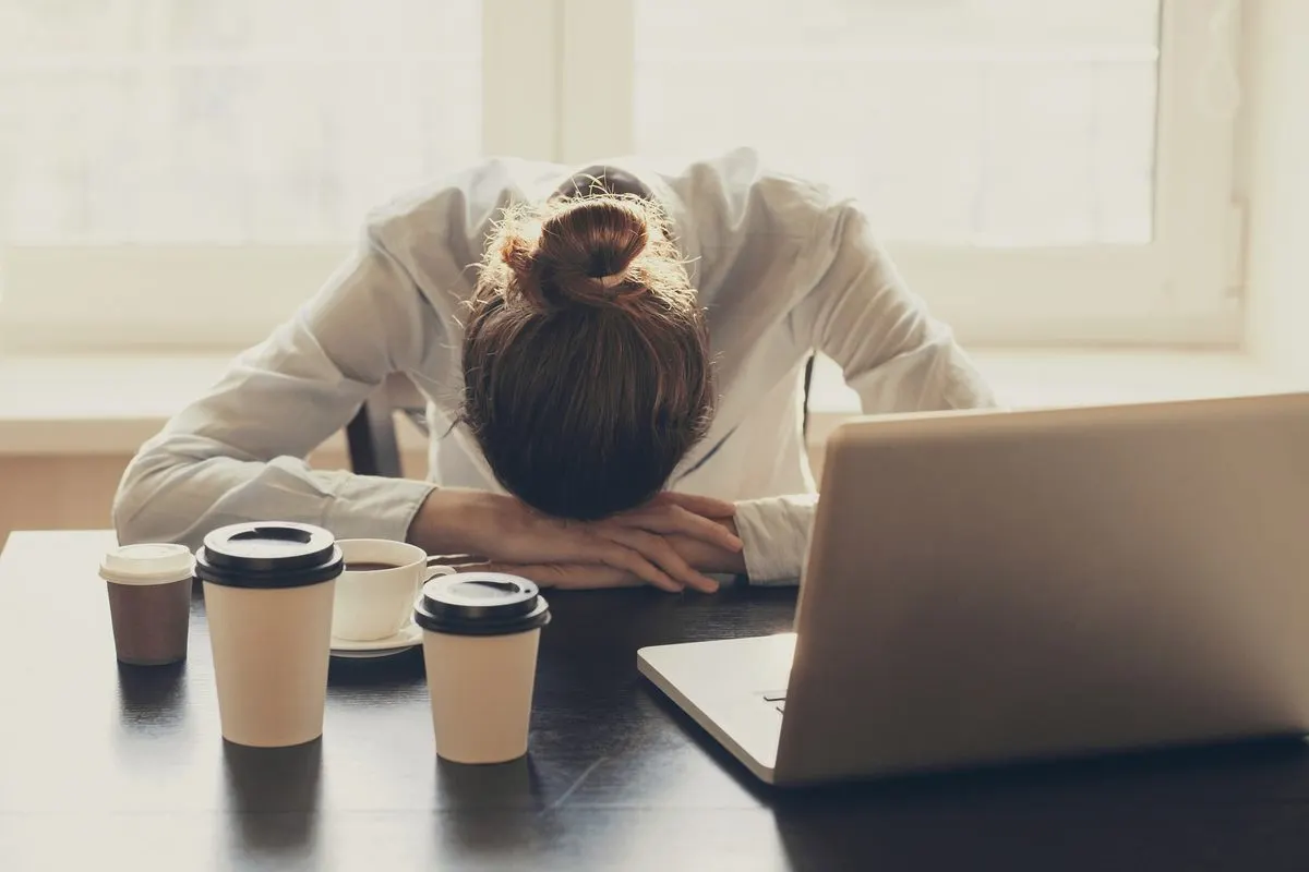 Eine Person ruht mit dem Kopf auf den Armen an einem dunklen Tisch, umgeben von Laptop und Kaffeetassen, vor einem hellen Fenster.