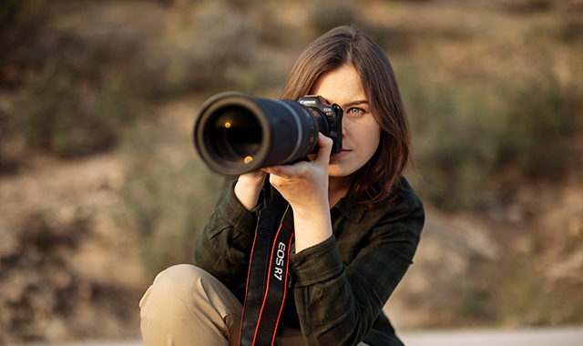 Eine Frau mit Brille hält eine Kamera mit großem Objektiv vor ihr Gesicht. Sie trägt eine Jacke und eine Kameragurt.