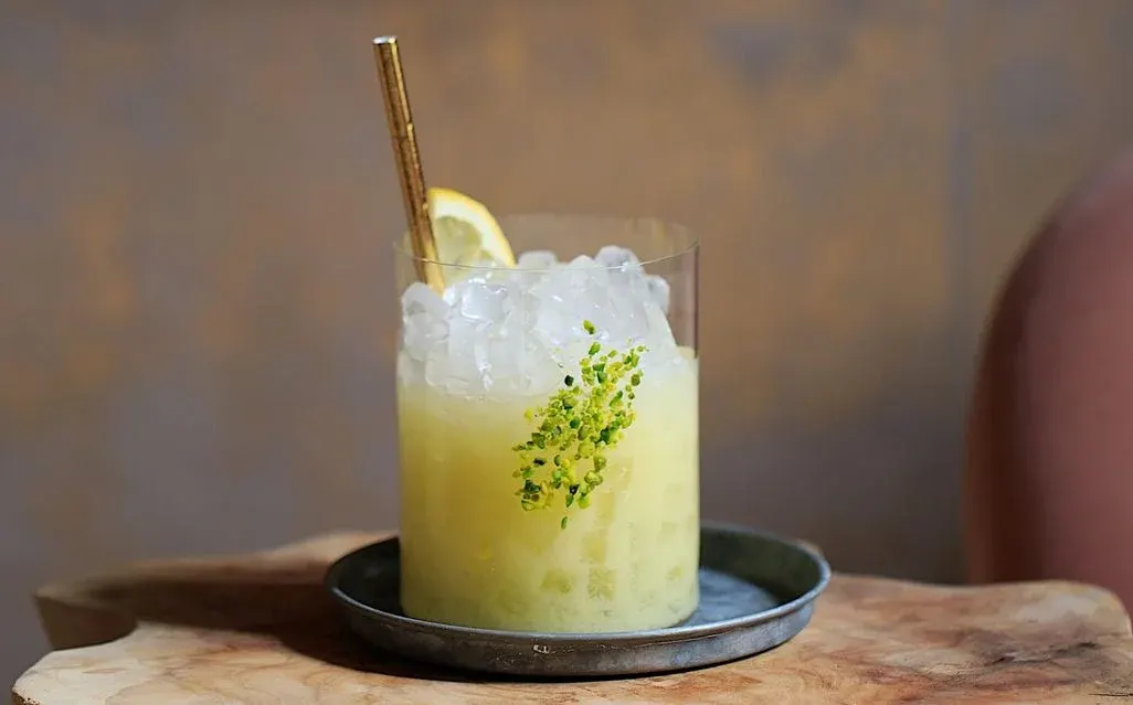 Glas mit gelbem Cocktail, zerstoßenem Eis, Zitronenscheibe, grüner Kräutergarnitur und goldenem Strohhalm auf Untersetzer.