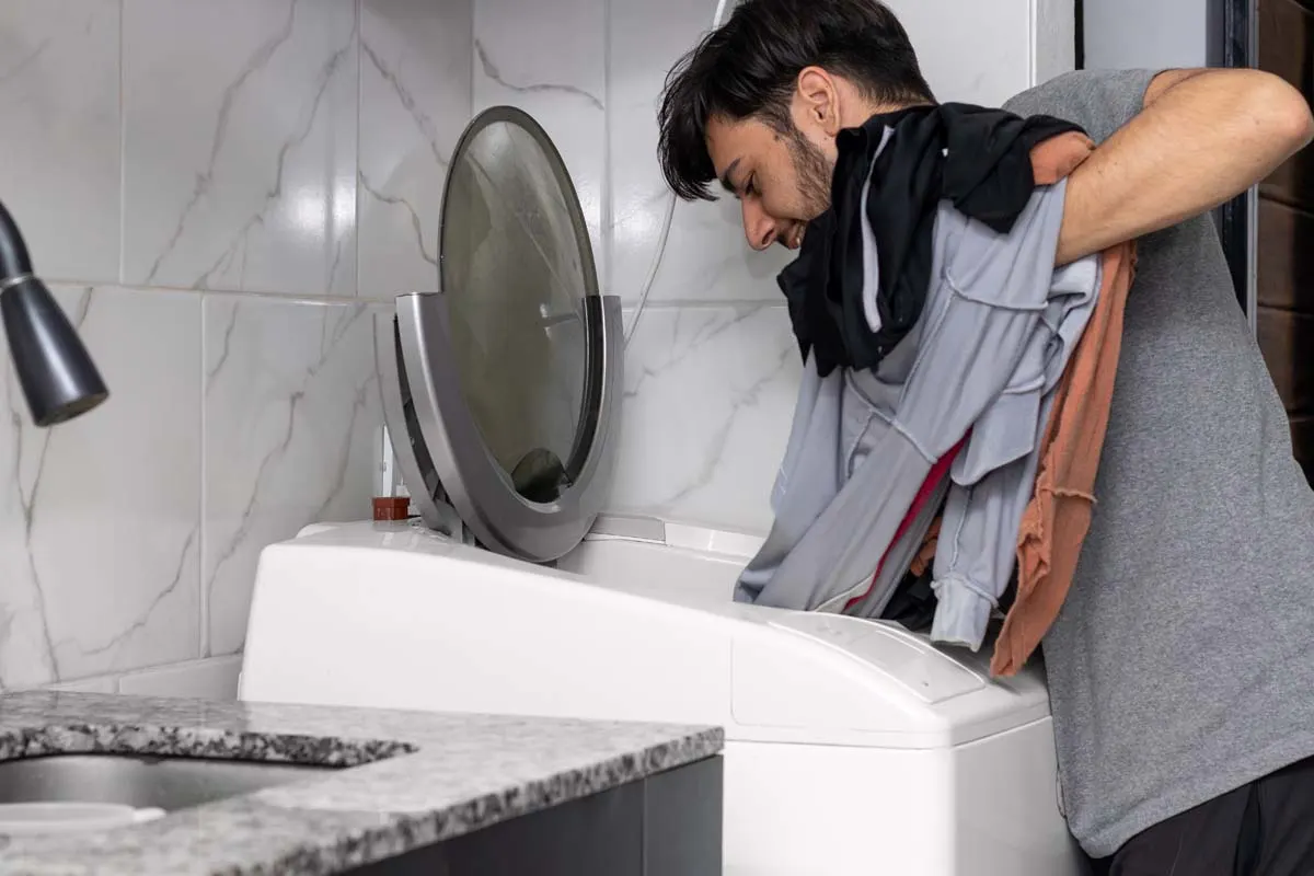 Mann mit dunklen Haaren beugt sich über eine geöffnete Toplader-Waschmaschine und legt Sportkleidung in Grau, Schwarz und Orange hinein, im Hintergrund weiss-graue Marmorfliesen und ein Waschbecken.