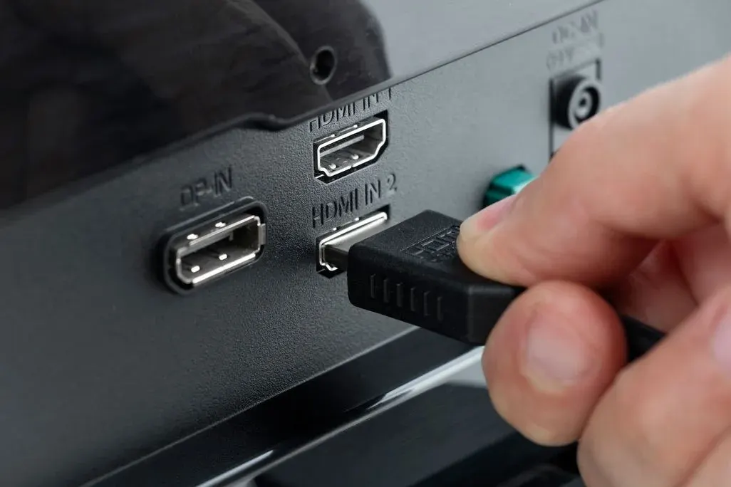 Eine Hand steckt ein schwarzes HDMI-Kabel in den HDMI IN 2-Anschluss eines schwarzen Geräts mit DP-IN und HDMI IN 1 Ports.