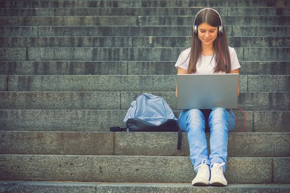 Sitzende Person auf Steinstufen nutzt silbernen Laptop, trägt weiße Kopfhörer mit rotem Kabel. Weißes T-Shirt, Jeans, weiße Sneaker. Grauer Rucksack daneben.