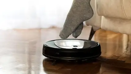 Person mit grauen Socken lässt die Füße spielerisch auf einem fahrenden Saugroboter über einen glänzenden Holzboden neben dem Sofa ruhen.