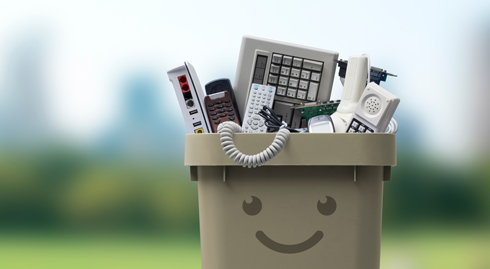 Ein beiger Behälter mit einem lächelnden Gesicht, gefüllt mit Elektronikschrott wie Tastaturen und Telefonen. Hintergrund ist unscharf.