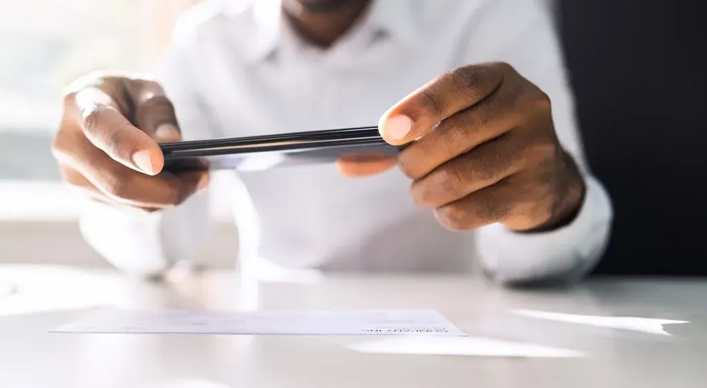 Eine Person in weißem Hemd fotografiert mit einem schwarzen Smartphone ein weißes Dokument auf einem hellen Tisch. Heller Hintergrund links, dunkel rechts.