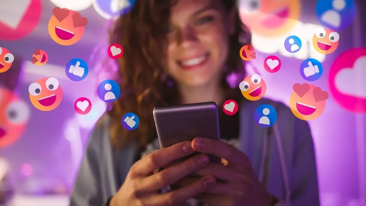 Person haelt ein Smartphone in den Haenden, umgeben von bunten Social-Media-Symbolen wie Herzen, Daumen hoch und lachenden Emojis.