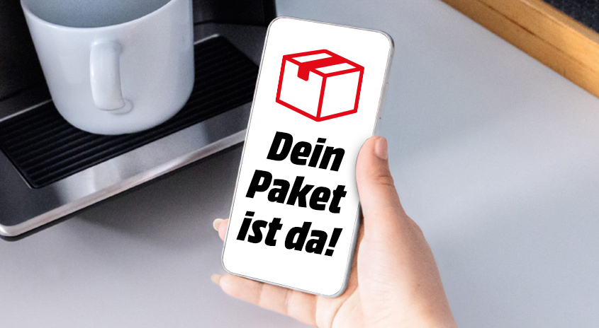 Smartphone in einer Hand zeigt weiße Bildschirmoberfläche mit rotem Paket-Icon und schwarzem Text ‚Dein Paket ist da!' neben einer weißen Kaffeetasse im Hintergrund.
