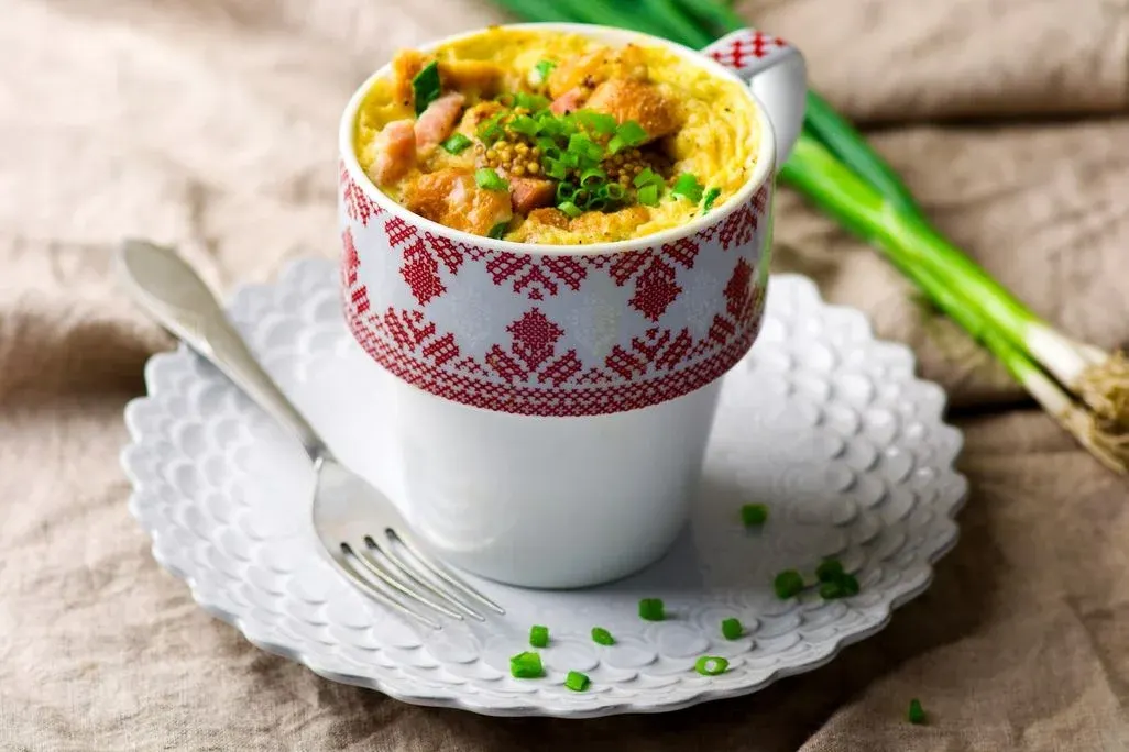 Eierspeise mit Croutons, Schinken, Senf und Lauchzwiebeln in weißer Tasse mit rotem Muster. Auf Unterteller, Gabel, beigem Tuch, Lauchzwiebeln.