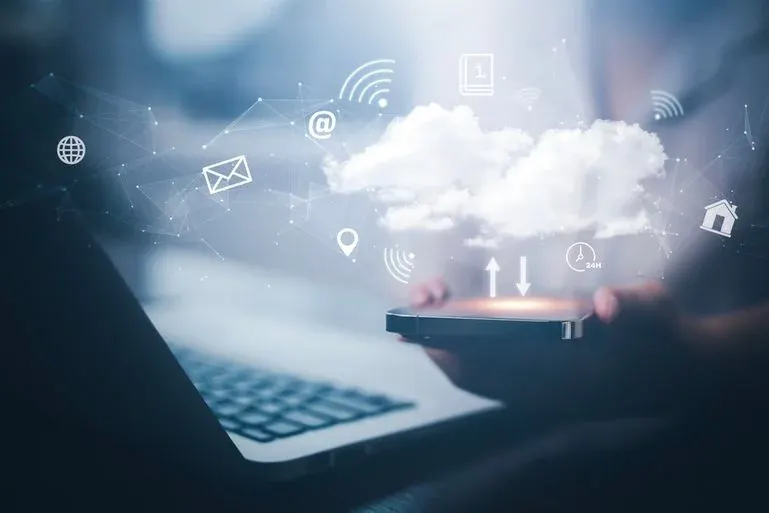 Hände halten ein Smartphone neben einem Laptop, umgeben von einer digitalen Wolke mit Netzwerk- und Anwendungssymbolen.