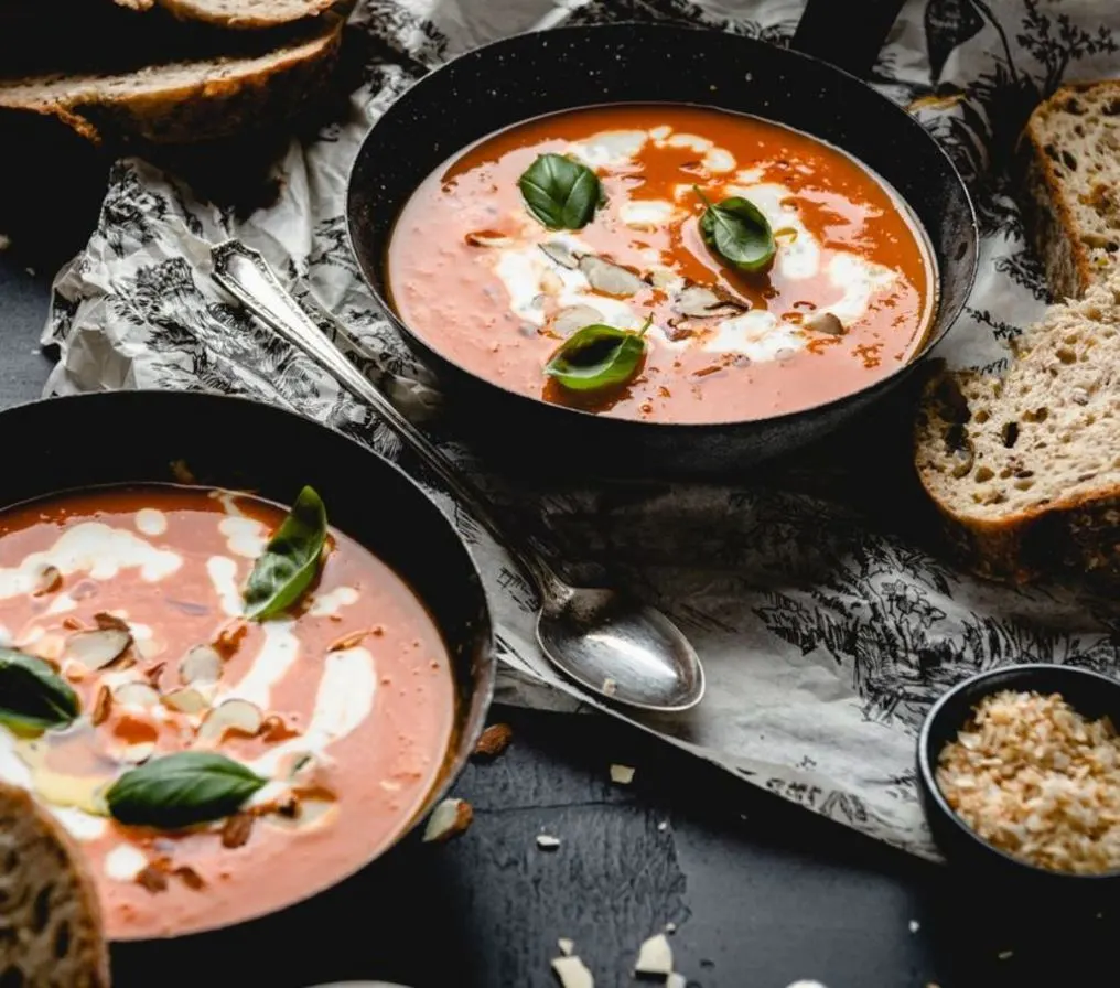 Zwei Schalen Tomatensuppe mit Obers, Basilikum und Brotscheiben daneben.
