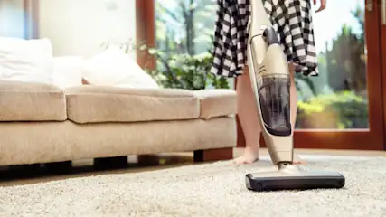 Person in kariertem Kleid saugt mit einem Akku-Staubsauger einen hellen Teppich vor einem Sofa.