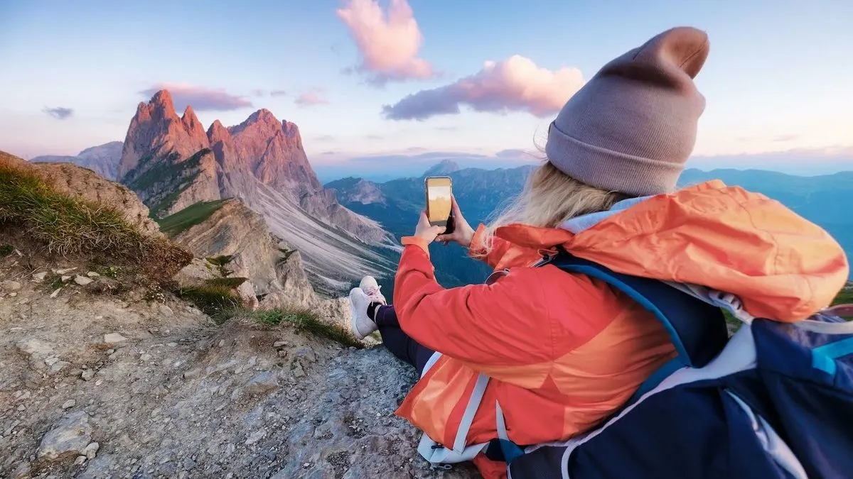 Frau sitzt auf einem Berg und fotografiert die Landschaft mit ihrem Smartphone.
