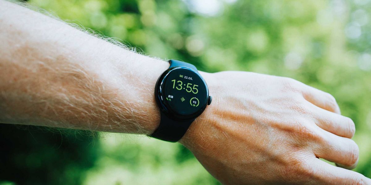 Eine Hand trägt eine schwarze Google Pixel Watch 2 mit schwarzem Display, das in grüner Schrift Datum, Uhrzeit und Fitnessdaten zeigt. Grüner Hintergrund.