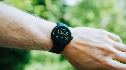 Eine Hand trägt eine schwarze Google Pixel Watch 2 mit schwarzem Display, das in grüner Schrift Datum, Uhrzeit und Fitnessdaten zeigt. Grüner Hintergrund.