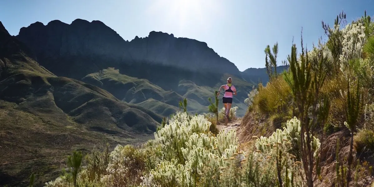 Frau in Sportkleidung joggt auf einem schmalen Pfad durch eine bergige Landschaft.
