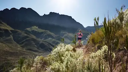 Frau in Sportkleidung joggt auf einem schmalen Pfad durch eine bergige Landschaft.