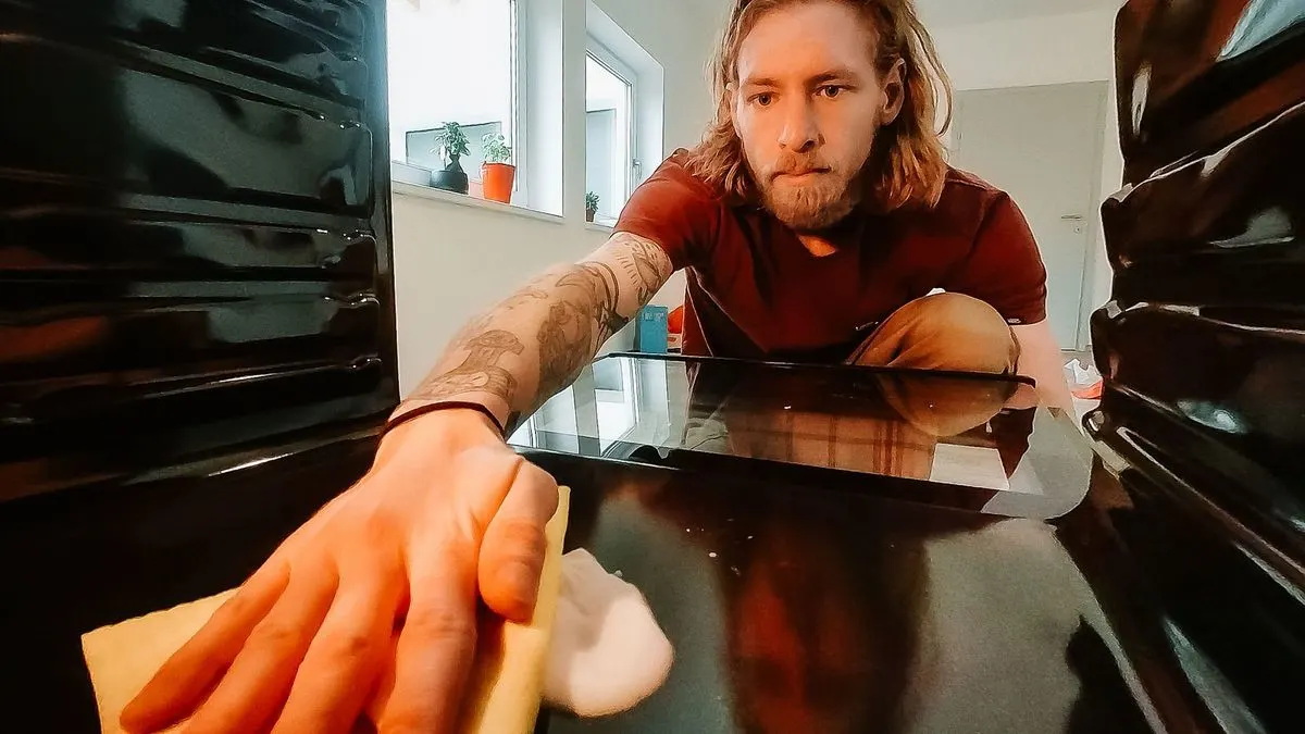 Eine Person mit tätowiertem Arm putzt eine dunkle Ofenoberfläche mit gelbem Tuch und Schaum. Im Hintergrund sind Fenster und Topfpflanzen.