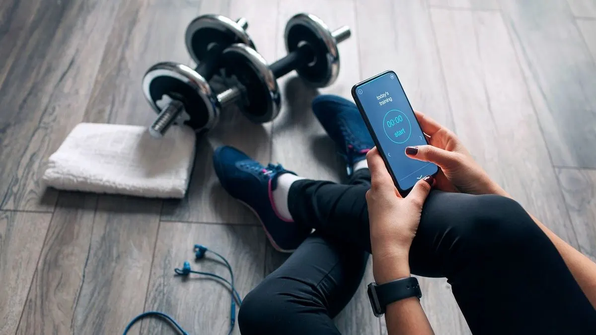 Person sitzt auf Holzboden in schwarzer Leggings und blauen Sportschuhen, hält Smartphone mit Fitness-App. Daneben Hanteln, weißes Handtuch und blaue Kopfhörer.