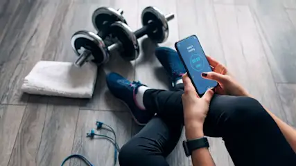 Person sitzt auf Holzboden in schwarzer Leggings und blauen Sportschuhen, hält Smartphone mit Fitness-App. Daneben Hanteln, weißes Handtuch und blaue Kopfhörer.