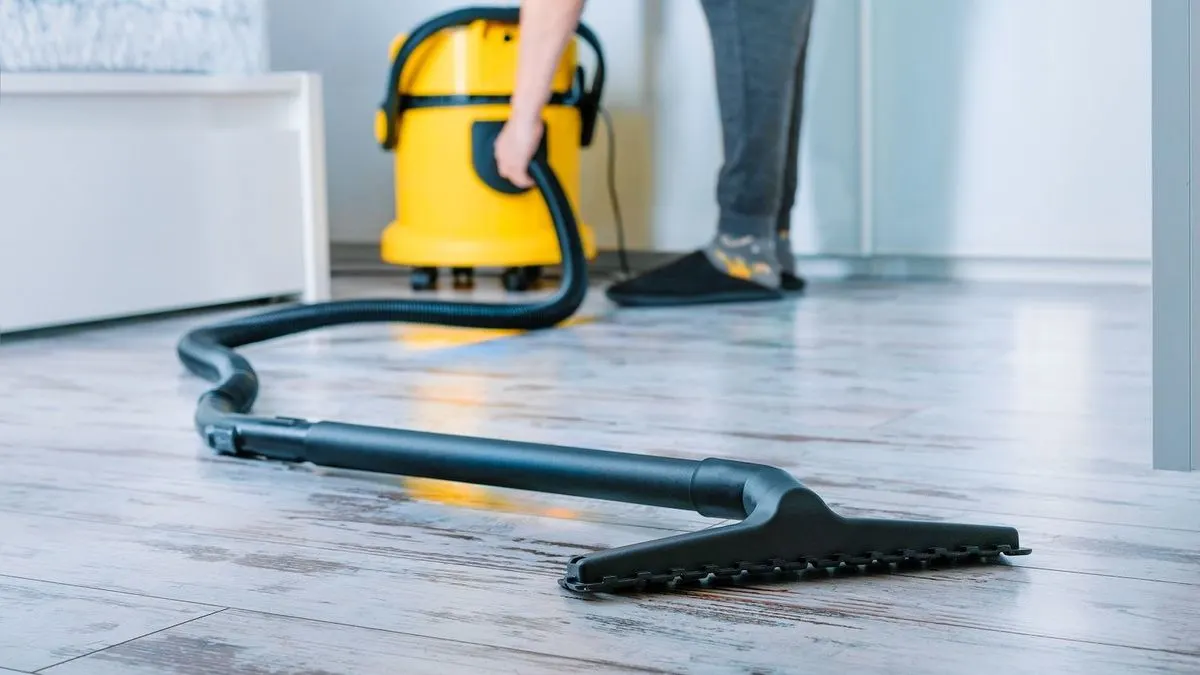 Gelber Staubsauger mit schwarzem Schlauch und Düse auf hellem Holzboden. Eine Person bedient den Schlauch vor weißem Möbel.