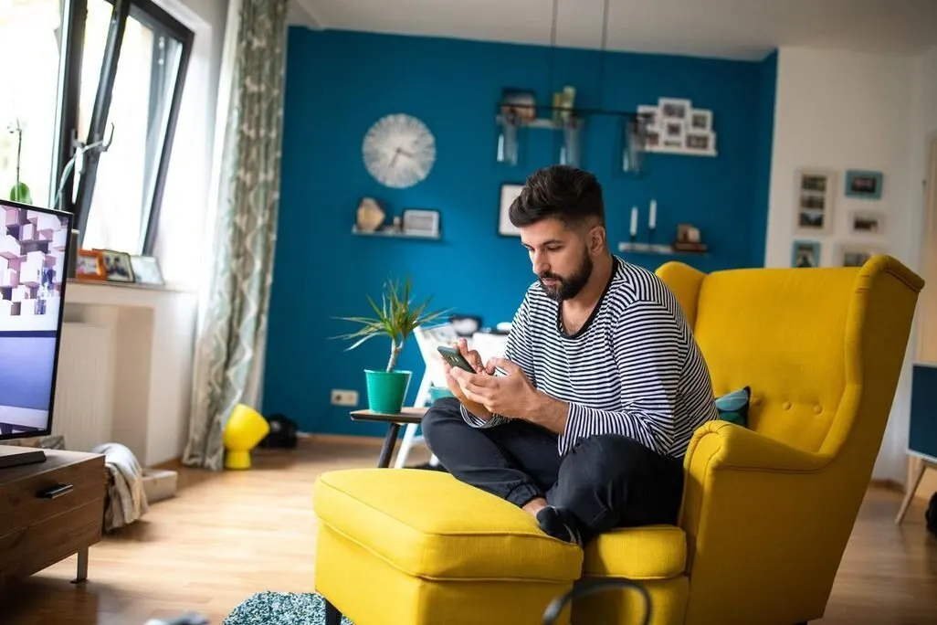 Mann sitzt in einem gelben Sessel und nutzt sein Smartphone im Wohnzimmer