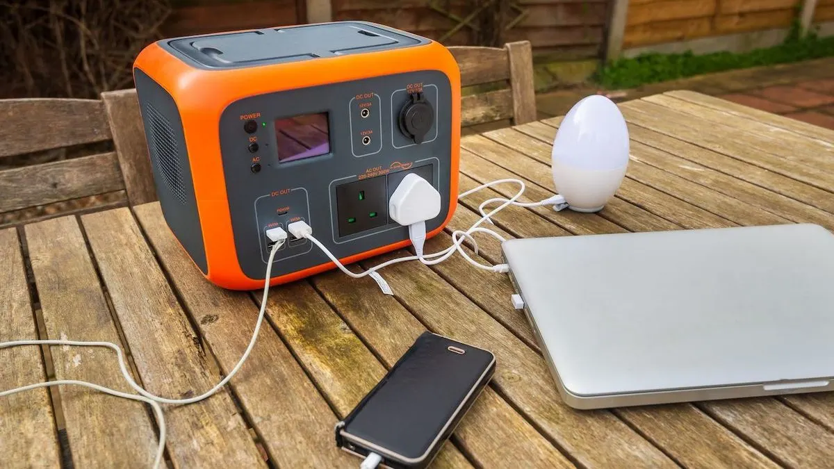 Eine orange-graue Powerstation auf einem Holztisch lädt ein silbernes Laptop, ein schwarzes Smartphone und eine weiße Lampe. Im Hintergrund sind ein Holzzaun und Grün zu sehen.