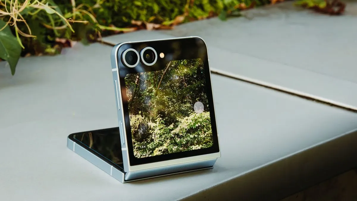 Silbernes faltbares Smartphone mit zwei Kameras auf hellgrauer Oberfläche. Der Bildschirm zeigt eine grüne Waldansicht. Unscharfes Grün im Hintergrund.