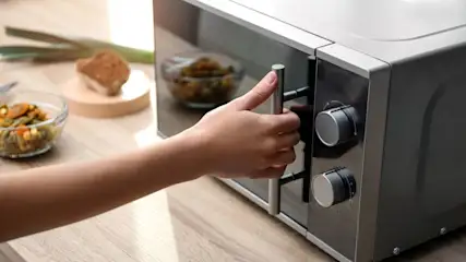 Eine geschlossene Mikrowelle steht auf einem Holztisch, eine Hand ist am Griff, im Hintergrund steht eine Schüssel mit Gemüse