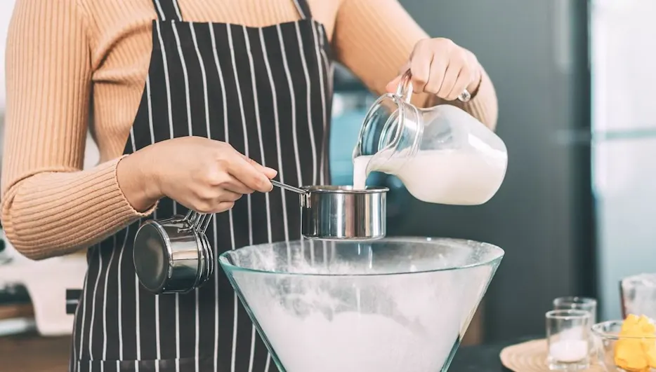 Person mit Schürze gießt Milch in einen Messbecher über einer großen Schüssel.