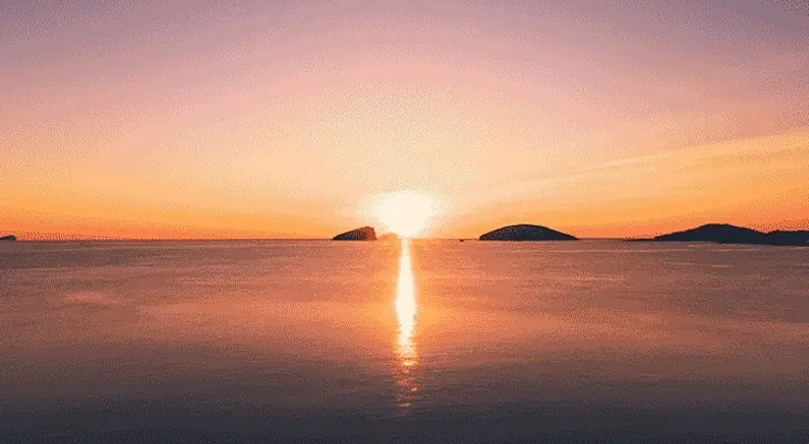 Sonnenaufgang über dem Meer mit mehreren kleinen Inseln am Horizont. Die Sonne spiegelt sich als heller Streifen im ruhigen Wasser, der Himmel leuchtet in warmen Orange- und Lilatönen.