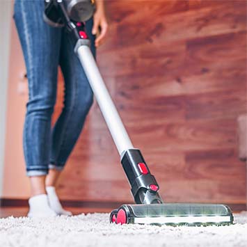 Staubsauger auf weißem Teppich vor Holzwand. Gerät ist silberfarben und schwarz mit roten Akzenten.