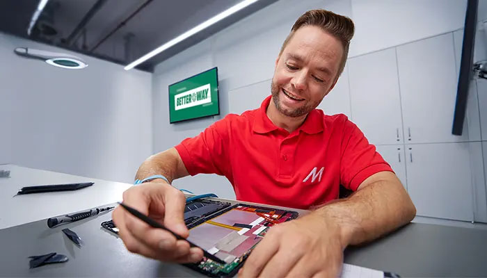 Ein Mann in rotem Hemd repariert ein elektronisches Gerät mit Werkzeugen auf einem Tisch in einem modernen Raum.