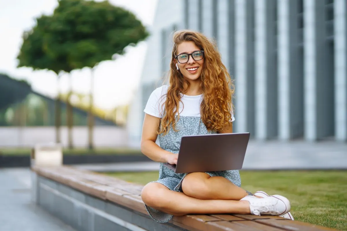 Eine blonde Studentin mit langen Haaren und Brille sitz fröhlich mit einem Laptop auf den Knien in Freien, im Hintergrund sieht man ein modernes Gebäude