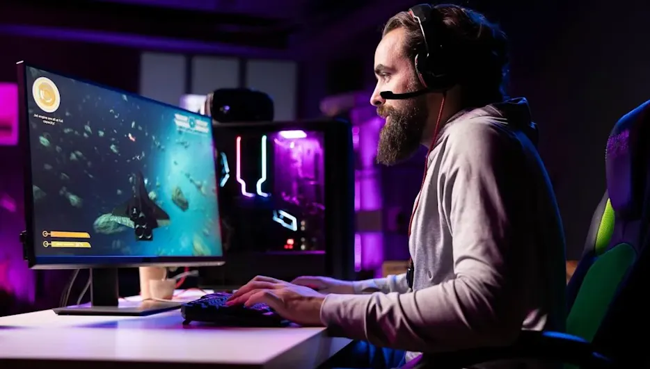Mann mit Headset sitzt an einem RGB-beleuchteten Gaming-PC und spielt konzentriert ein Weltraumspiel auf großem Monitor in abgedunkeltem Zimmer.