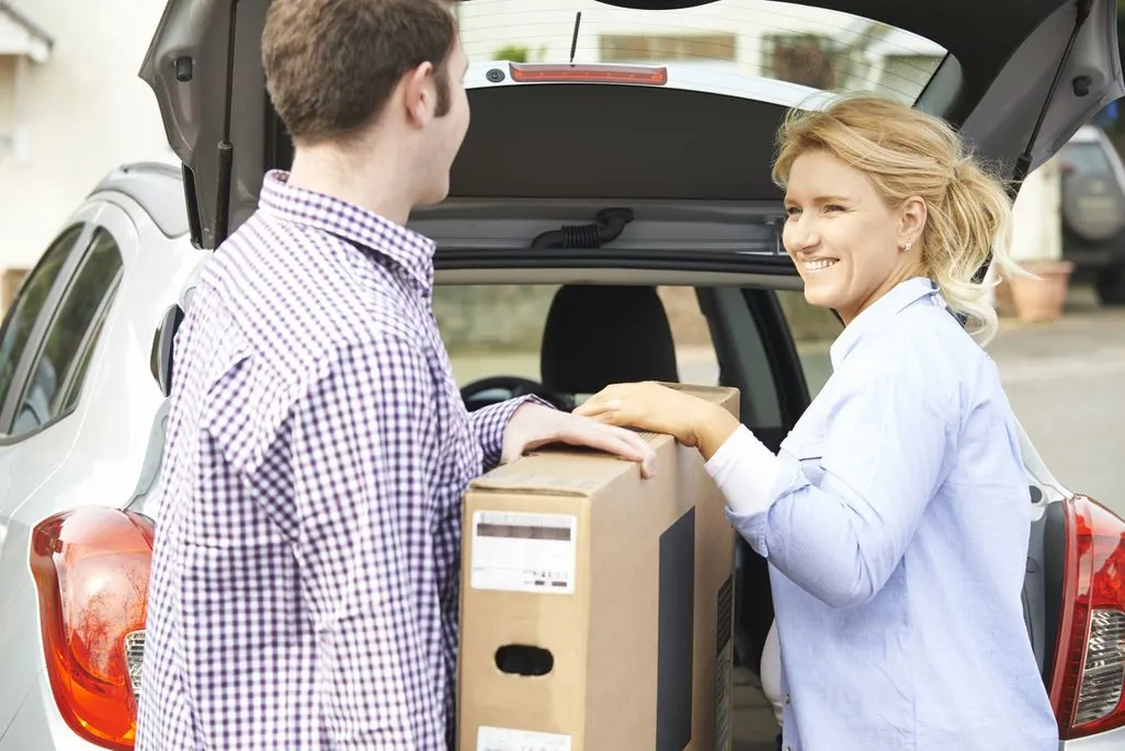 Zwei Personen laden gemeinsam einen großen Karton in den Kofferraum eines Autos.