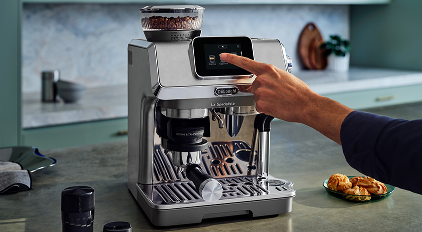 Eine silberne De'Longhi Kaffeemaschine auf einer Küchentheke, ein Finger berührt den Bildschirm. Im Hintergrund sind Küchenutensilien.