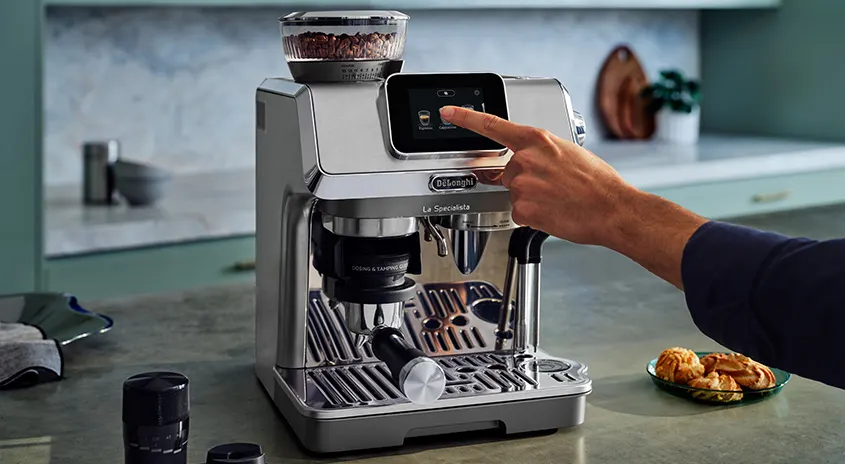 Eine silberne De'Longhi Kaffeemaschine auf einer Küchentheke, ein Finger berührt den Bildschirm. Im Hintergrund sind Küchenutensilien.