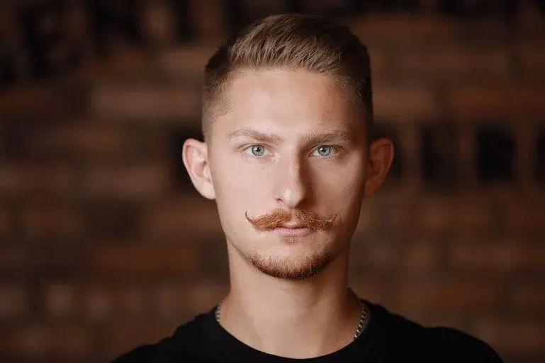 Ein Mann mit gezwirbeltem Schnurrbart, Kinnbart, hellem Blick und Kette trägt ein dunkles Shirt.