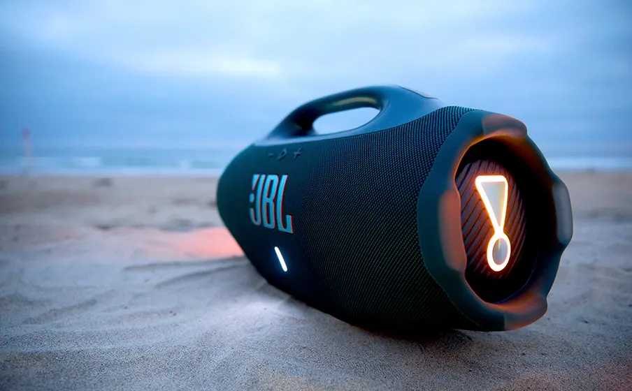 Schwarzer JBL-Bluetooth-Lautsprecher mit orangefarbener LED-Beleuchtung am Sandstrand vor bewölktem Himmel.