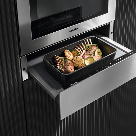 Ein Miele-Ofen mit einer schwarzen Form mit Lammkoteletts und Gemüse auf einem silbernen Auszug vor einer dunklen, geriffelten Wand.