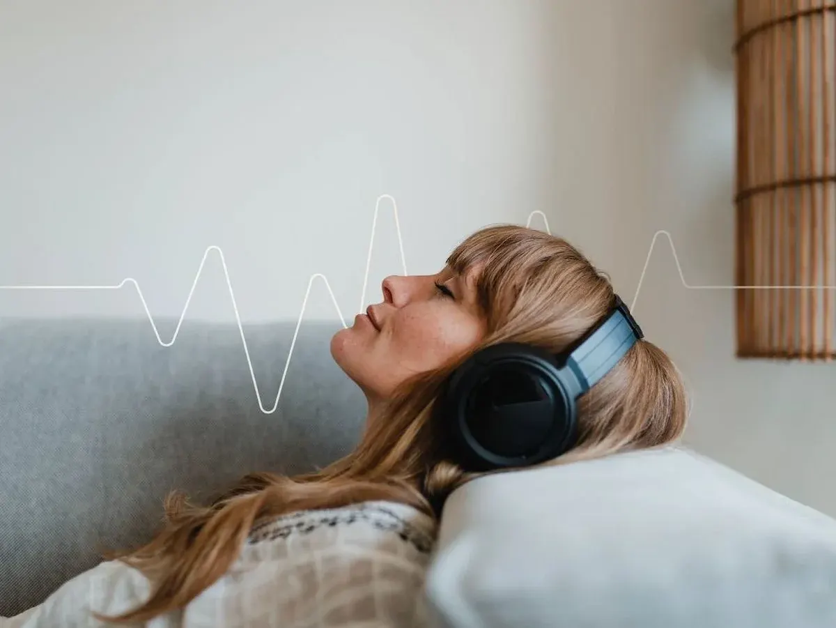 Frau hört Musik mit Over‑Ear‑Kopfhörern auf dem Sofa, entspannter Gesichtsausdruck, Frequenzgrafik im Hintergrund.