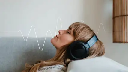 Frau hört Musik mit Over‑Ear‑Kopfhörern auf dem Sofa, entspannter Gesichtsausdruck, Frequenzgrafik im Hintergrund.