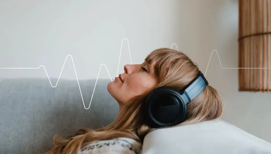 Frau hört Musik mit Over‑Ear‑Kopfhörern auf dem Sofa, entspannter Gesichtsausdruck, Frequenzgrafik im Hintergrund.