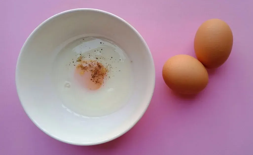Eine weiße Schale enthält ein weichgekochtes Ei mit Pfeffer und Gewürzen. Zwei rohe braune Eier liegen daneben auf einer pinken Oberfläche.
