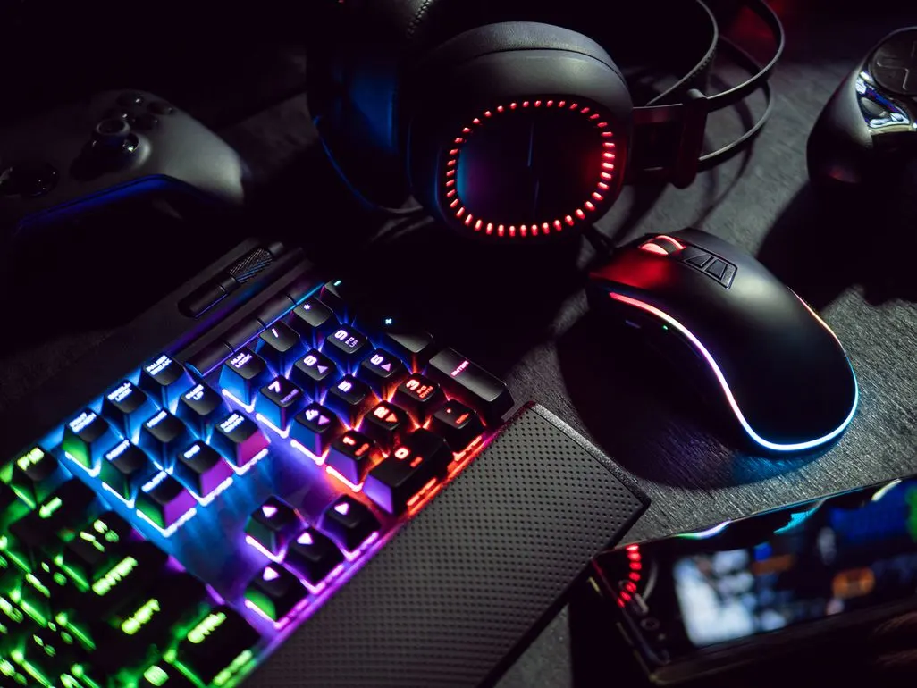 Gaming-Setup mit beleuchteter RGB-Tastatur, Maus und Kopfhörern auf dunkler Oberfläche, dazu Controller und Smartphone.