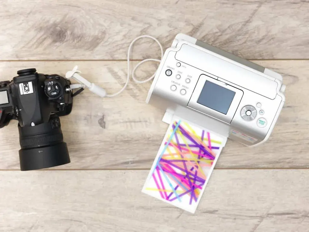 Schwarze digitale Spiegelreflexkamera mit Objektiv und weißem tragbarem Fotodrucker mit LCD-Display auf Holzuntergrund – der Drucker gibt gerade ein buntes Sofortfoto mit magentafarbenen und gelben Mustern aus, verbunden mit weißem USB-Kabel.