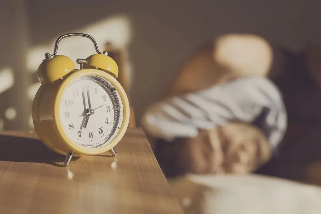 Gelber Wecker auf Holztisch zeigt 7 Uhr; im unscharfen Hintergrund schläft eine Person unter weißer Decke im Sonnenlicht.